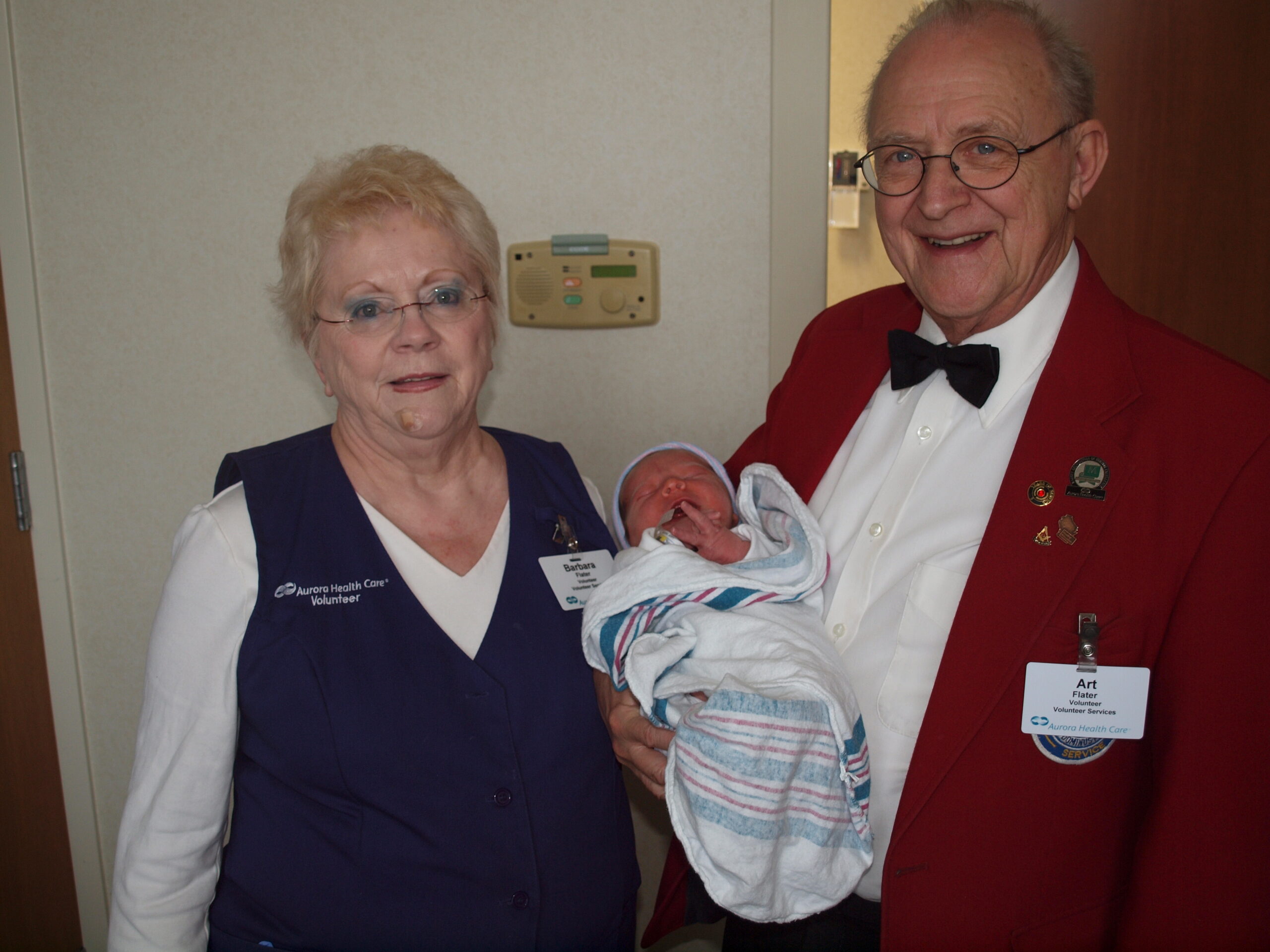 Two elderly adults pose together: a woman in a dark vest and a man in a red blazer holding a newborn wrapped in a blanket; both smiling in a hospital setting.
