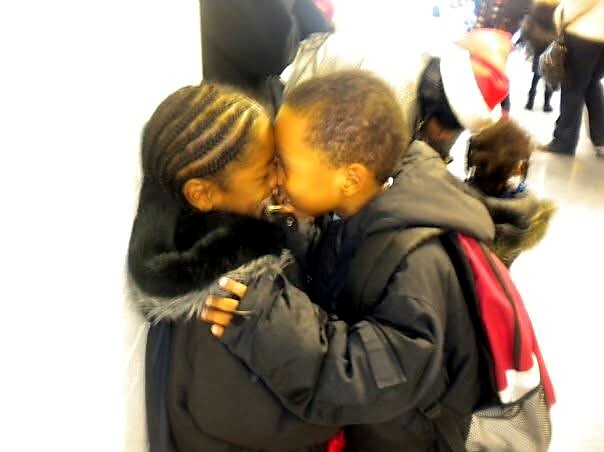 Two young children in winter jackets share a kiss in a hallway, a backpack visible on one child.