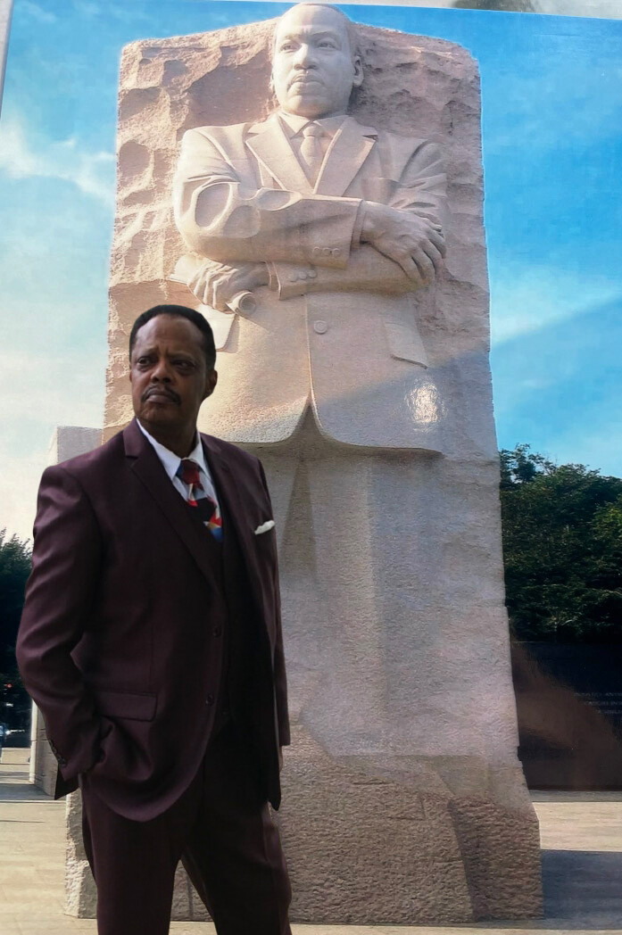 Man in a burgundy suit posing beside a large stone statue with arms crossed, outdoors on a sunny day.