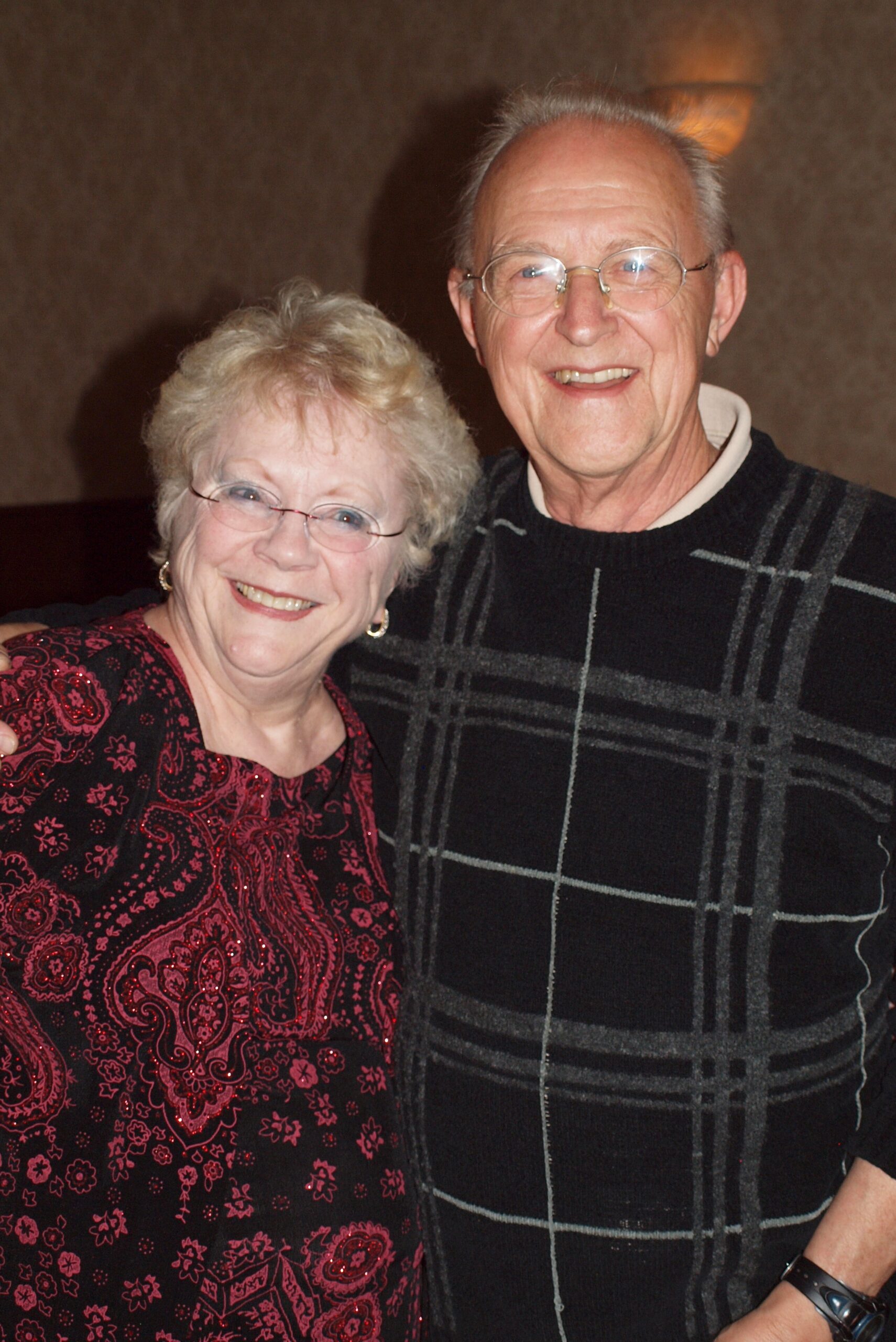 Smiling elderly couple standing close together indoors, woman in red patterned top and man in dark plaid sweater.