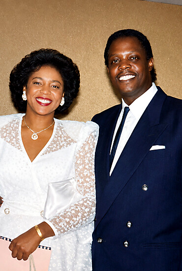 Smiling couple in formal attire posing for a photo: woman in a white lace dress, man in a navy suit with a tie.