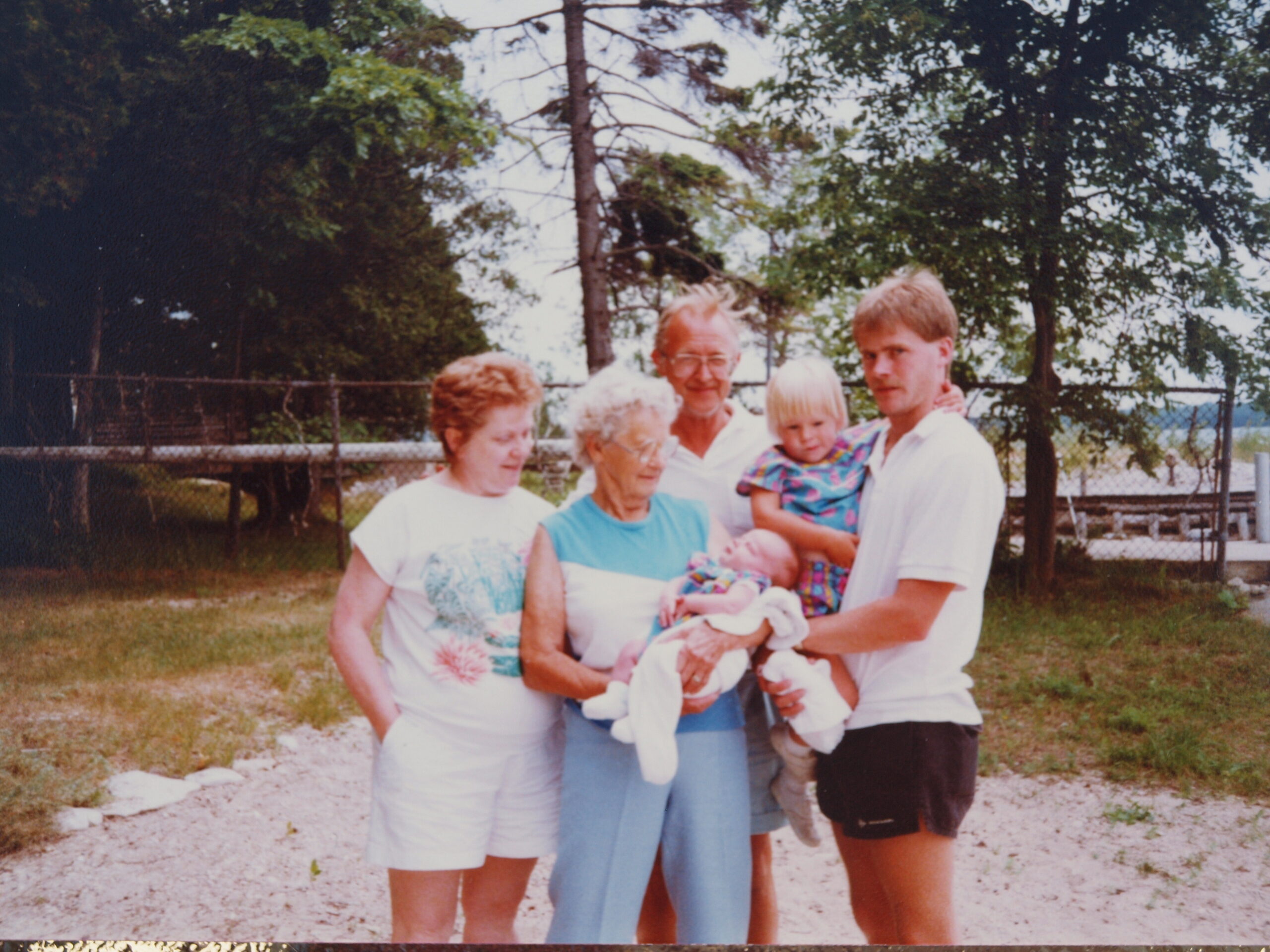 Family group posing outdoors in a yard, with an older woman holding a baby wrapped in a colorful blanket while others stand nearby.