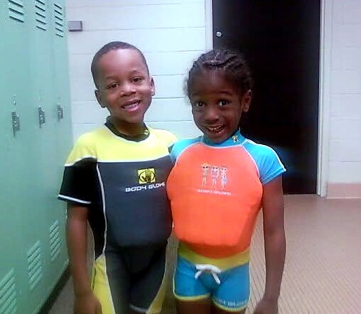 Two smiling children wearing colorful wetsuits in a school locker room, posing for the camera.