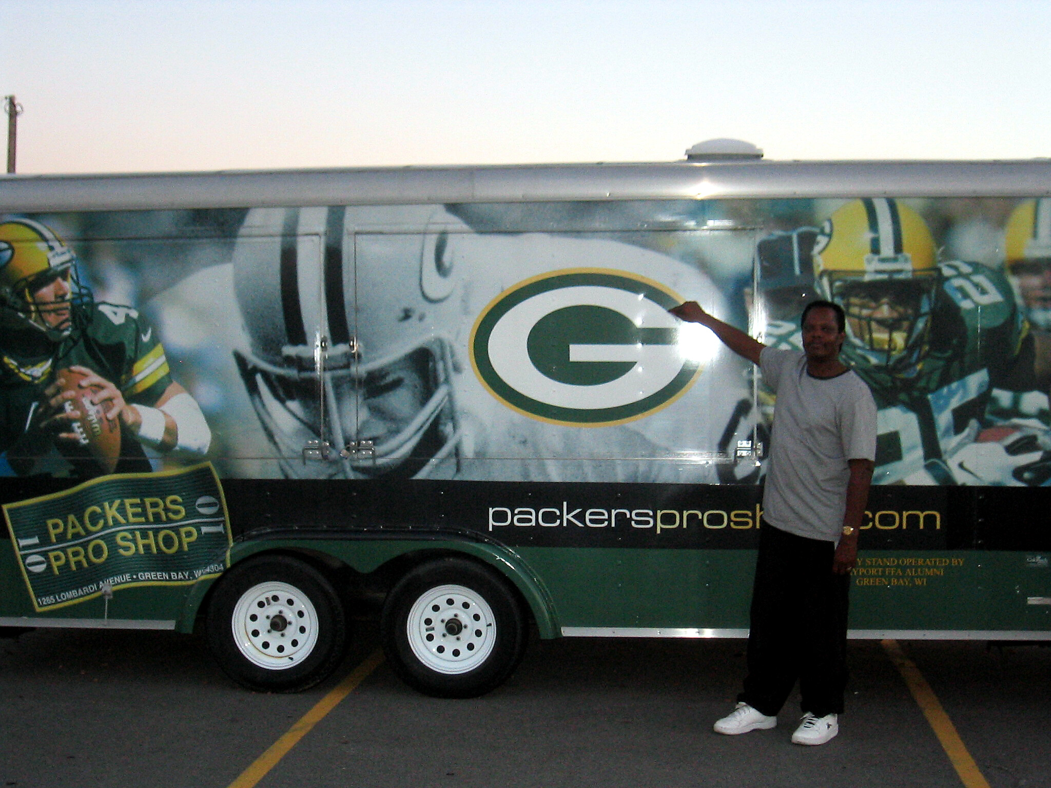 Green Bay Packers promotional trailer with large logo graphics; a man in a gray shirt stands in front in a parking lot beside the trailer.