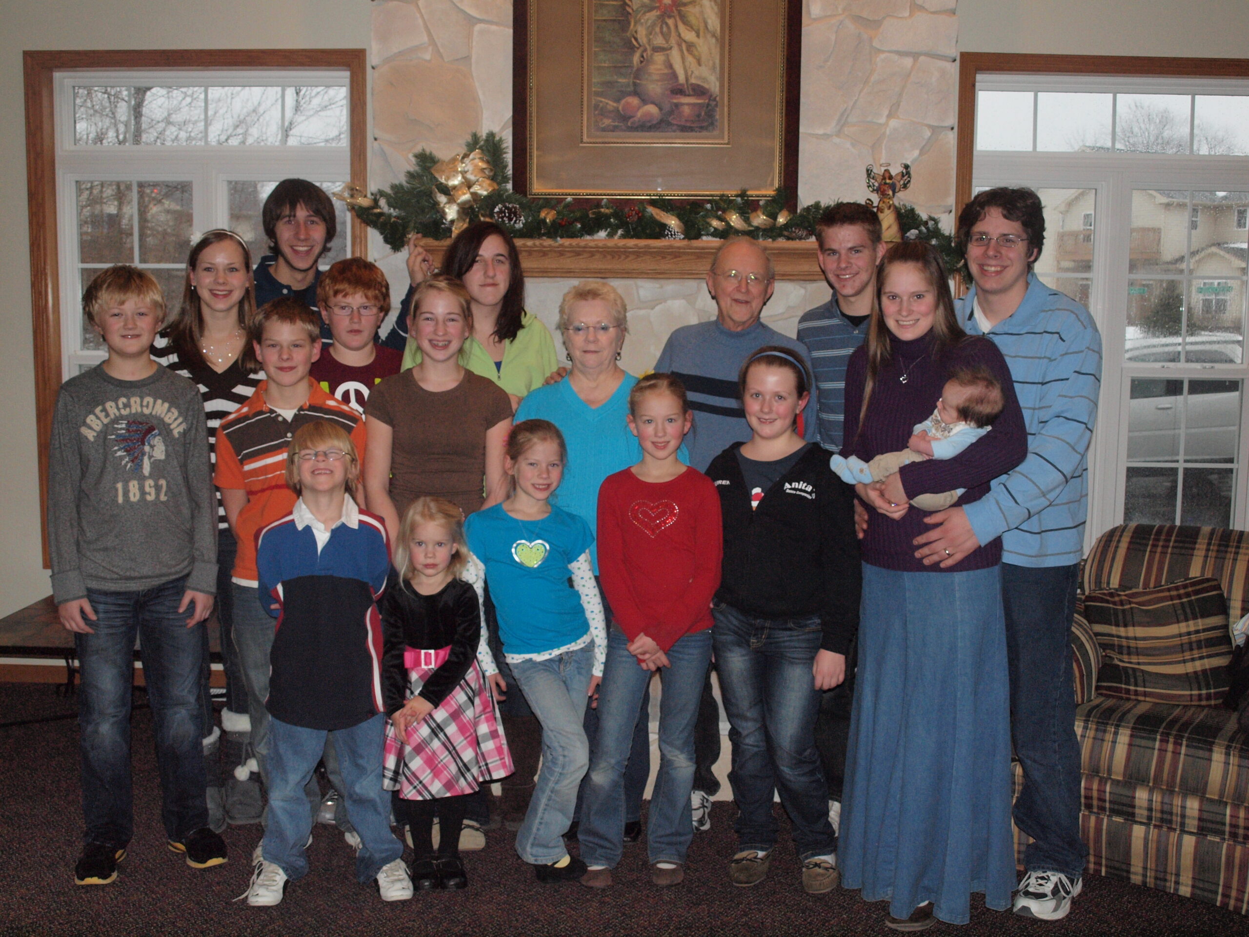 Group photo of many children and adults in a living room with a stone fireplace and holiday garland outside the frame windowed walls.