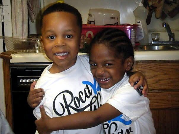 Two smiling children wearing matching white shirts hug in a kitchen, arms around each other.