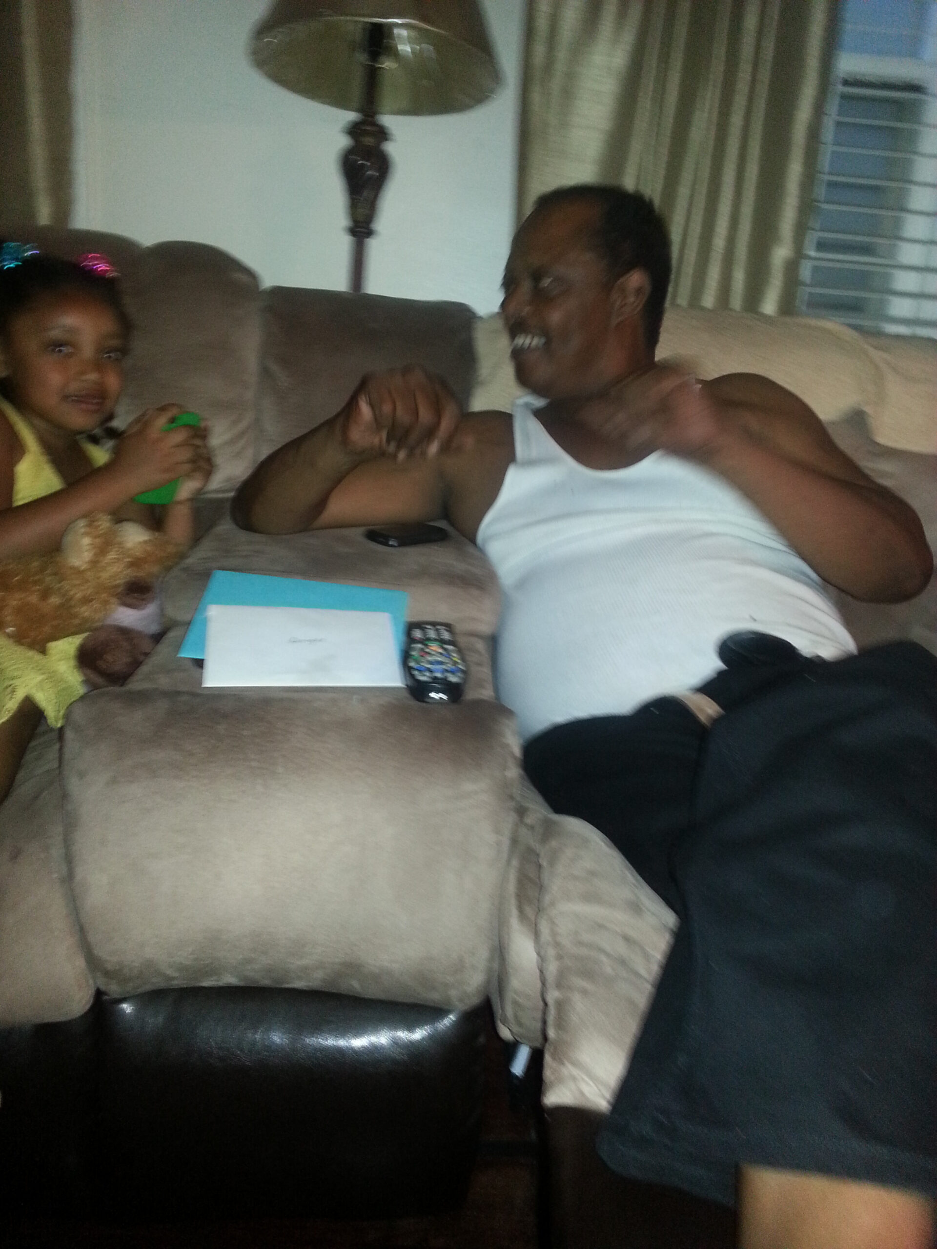 Man in a white tank top on a couch smiling while a young girl in a yellow dress sits beside him with a green cup and a stuffed animal nearby on the couch.