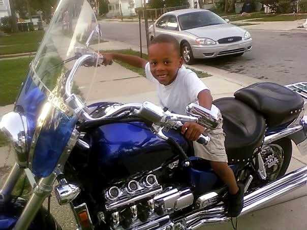 Young boy sits on a large blue motorcycle in a suburban driveway, smiling at the camera.