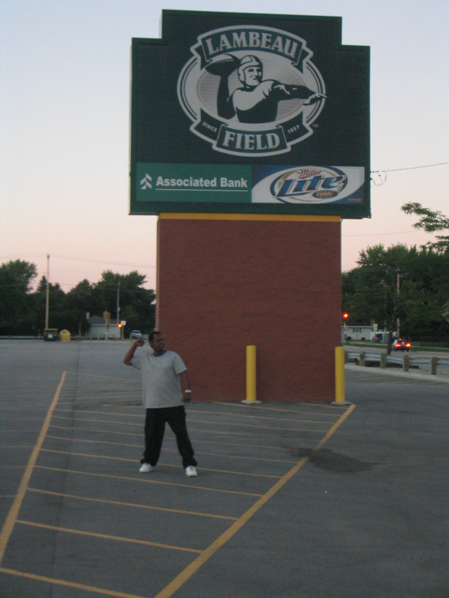 Man in a light gray shirt stands in a parking lot in front of a large Lambeau Field sign with Associated Bank and Miller Lite logos.