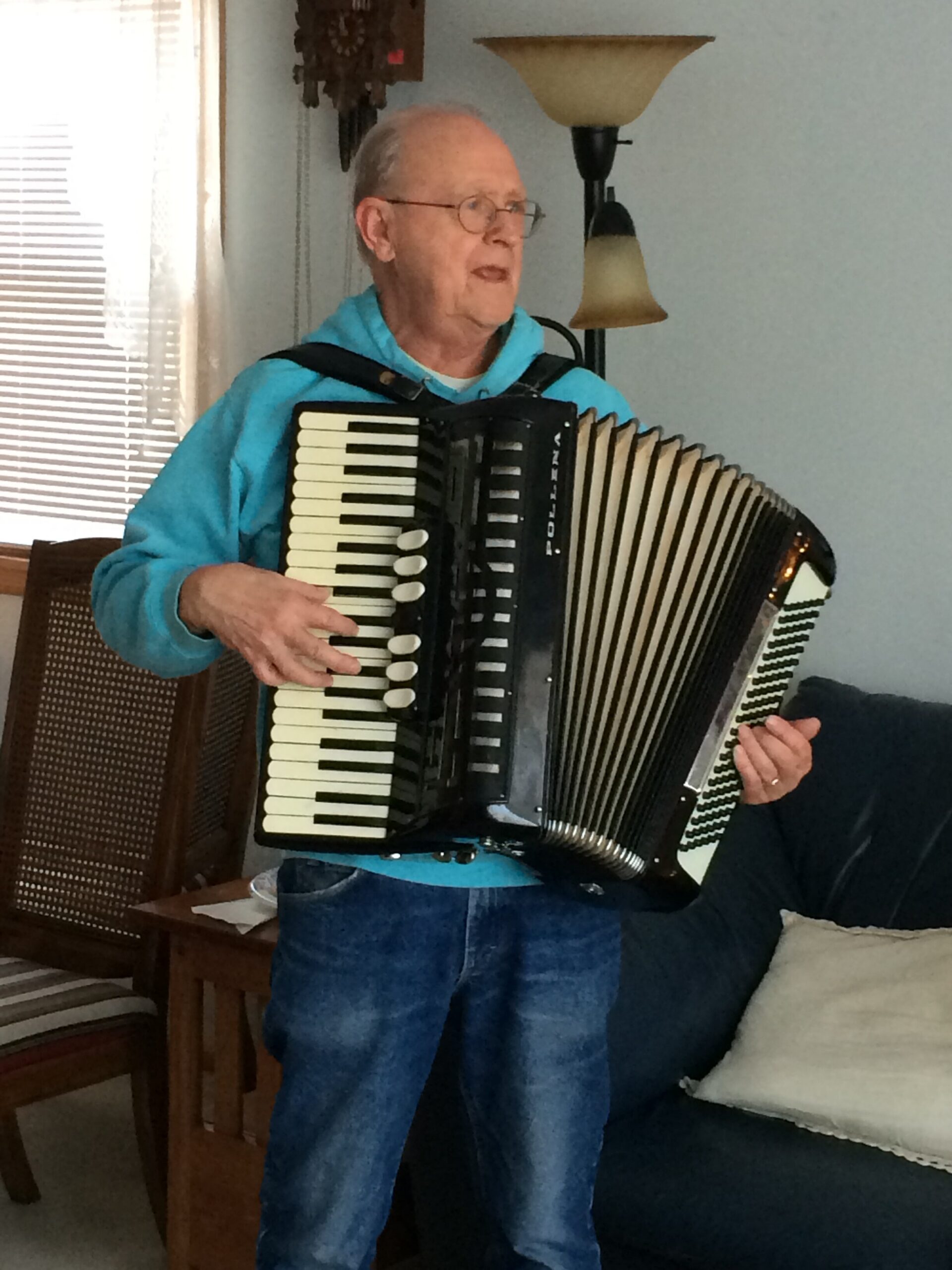 Older man wearing glasses and a blue jacket plays a large black-and-white accordion indoors in a living room setting.