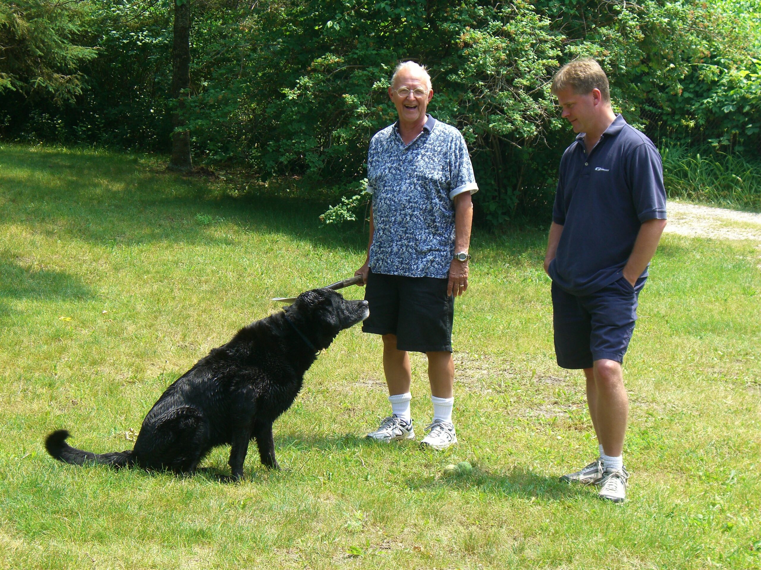 Two men stand in a sunlit grassy yard with a black dog on a leash; the older man smiles at the camera while the younger man looks down.