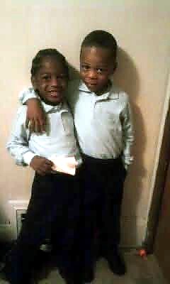 Two kids standing side by side in matching outfits, arms around each other; the younger child on the left holds a small envelope.