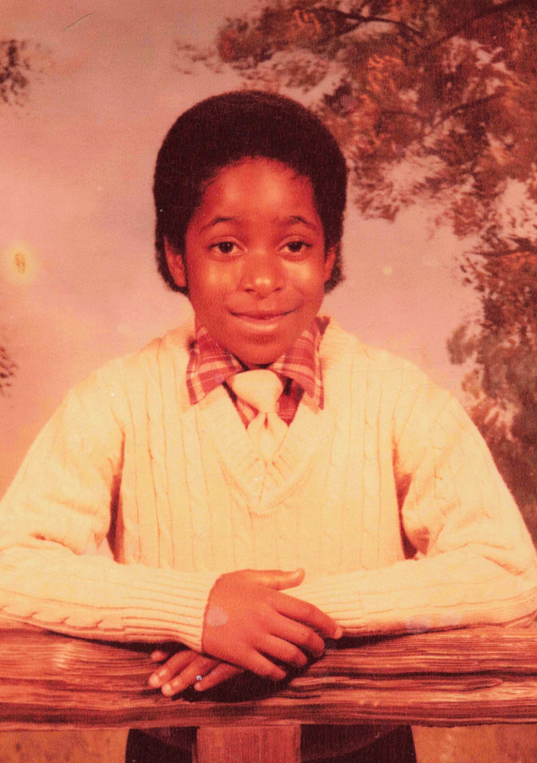 Young boy with an Afro hairstyle, wearing a light striped sweater and plaid shirt, sitting with hands folded on a wooden table.