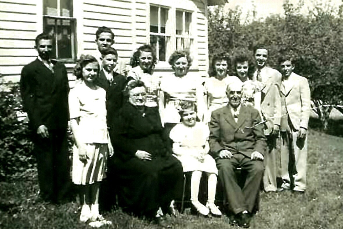 Multigenerational family group photo outdoors, with adults standing and two elderly people seated, in front of a wooden house and greenery.