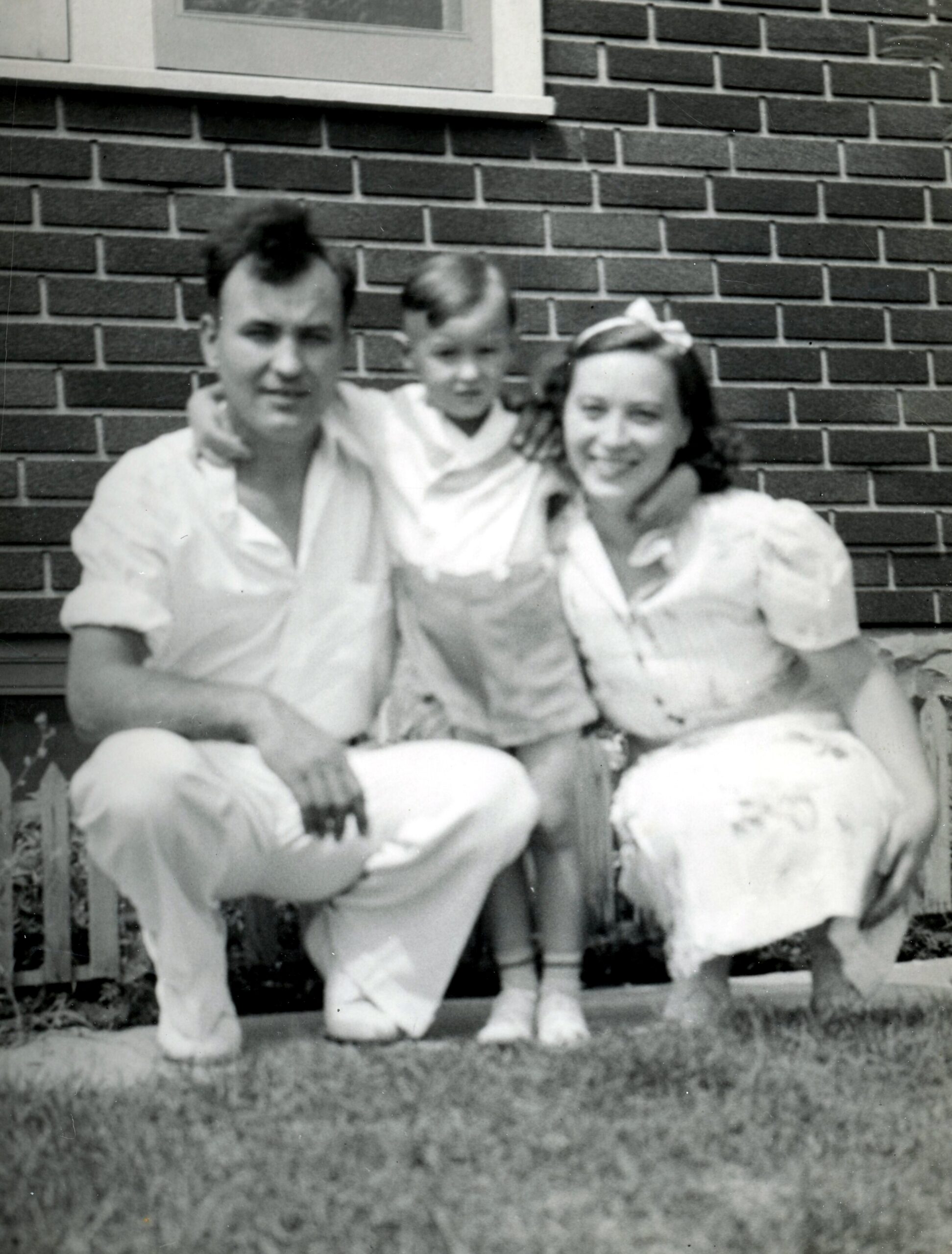 Black-and-white photo of a family of three posing on a porch in front of a brick wall: man on the left, child in the middle, woman on the right.