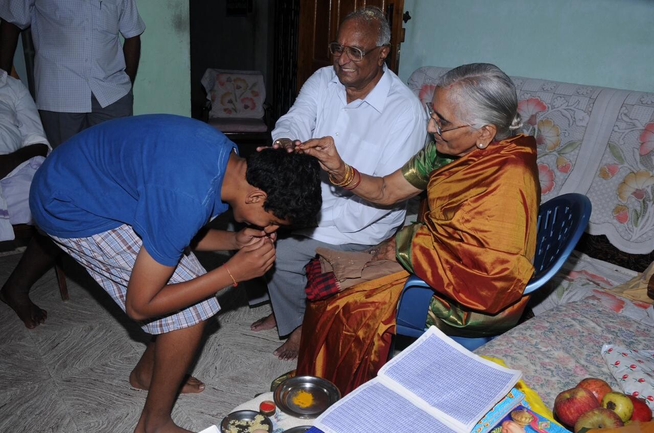 Elderly woman in an orange sari blessing a young man as he bows his head indoors while others sit nearby.