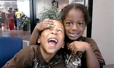 Two smiling girls in brown shirts pose for the camera; one playfully makes a goofy face while the other hugs her friend.
