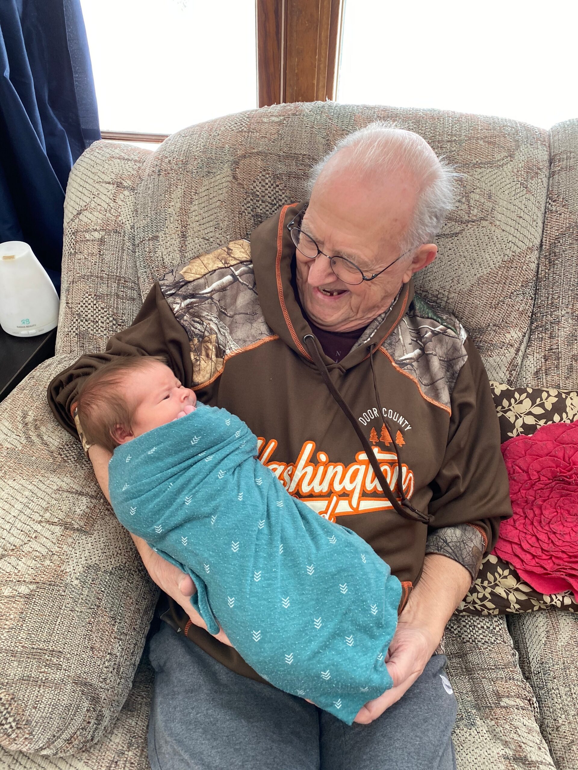 Older man with glasses sits in a patterned armchair, cradling a newborn wrapped in a teal blanket.