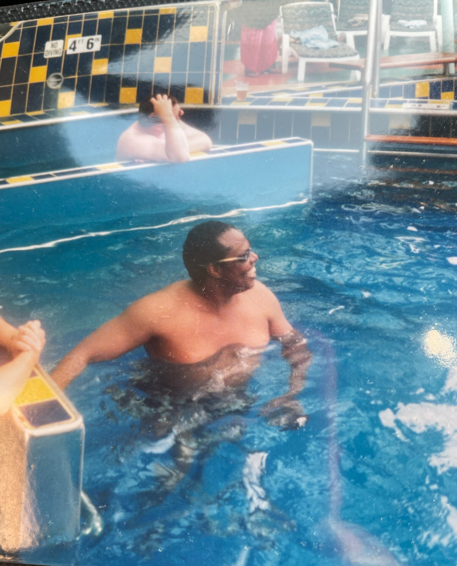 Two men in a swimming pool: the man in the foreground is shirtless with glasses, while another person near the pool wall holds his head with his hands beside a yellow-and-black tiled edge.