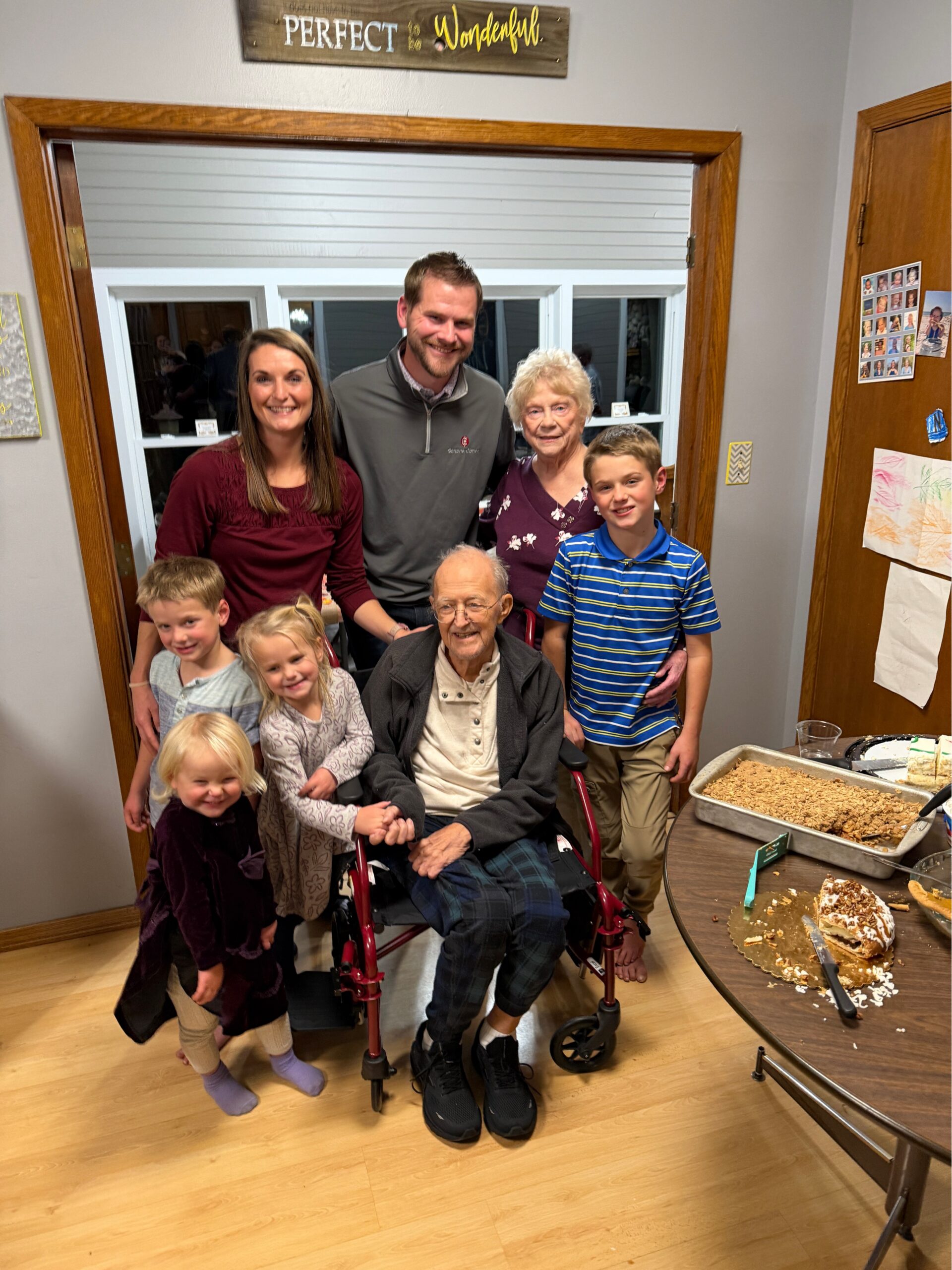 Smiling group of family members around an elderly man in a wheelchair, indoors with cake on the table and crumbs nearby.