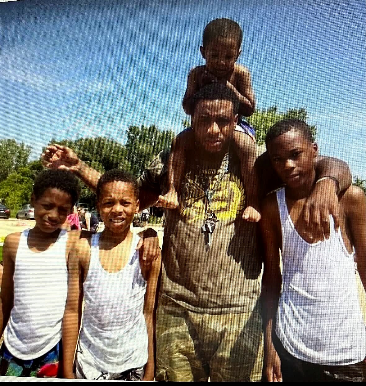 Adult man with four boys posing outdoors on a sunny day; a boy sits on the man's shoulders.