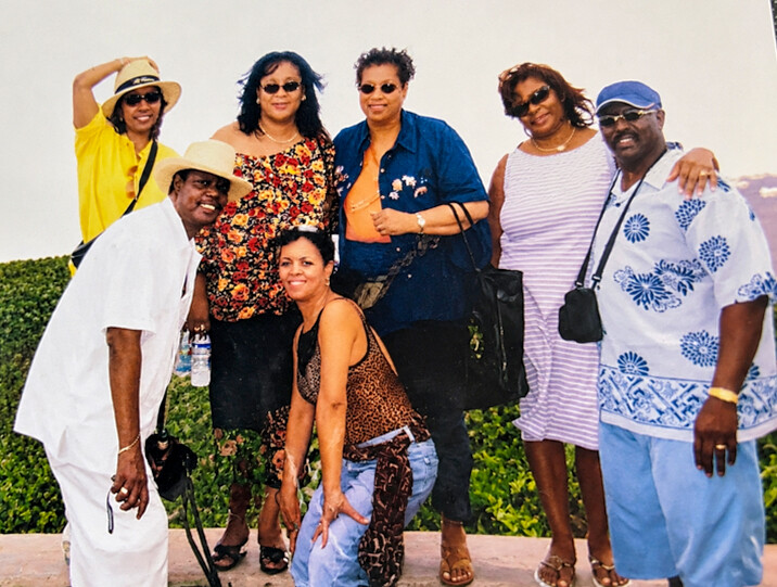 Group of seven adults posing for a casual outdoor photo on a sunny day, wearing hats, sunglasses, and summery outfits with greenery in the background.