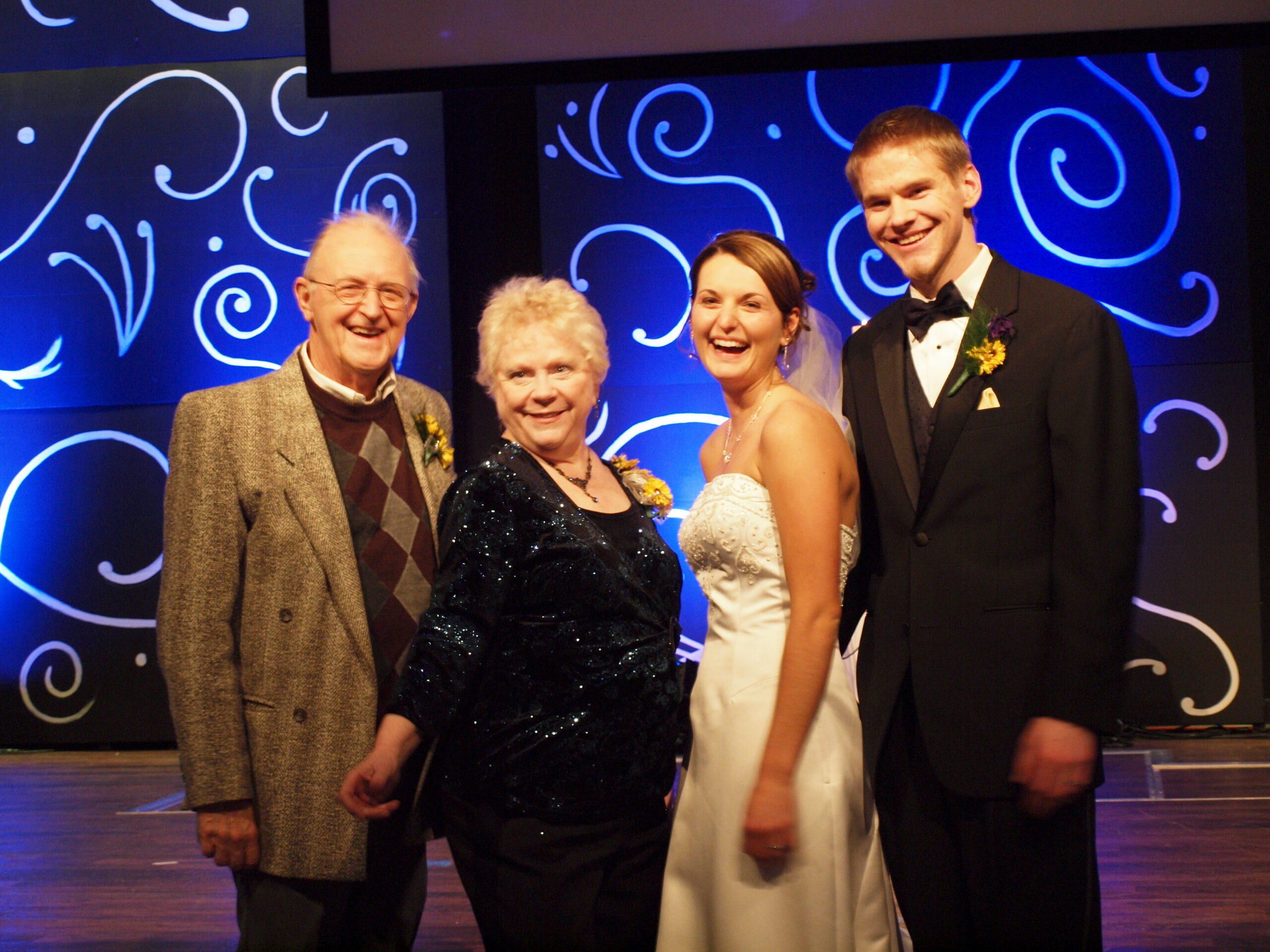 Four people in formal attire pose together on a stage with a blue decorative swirl backdrop, smiling for a group photo at an event.