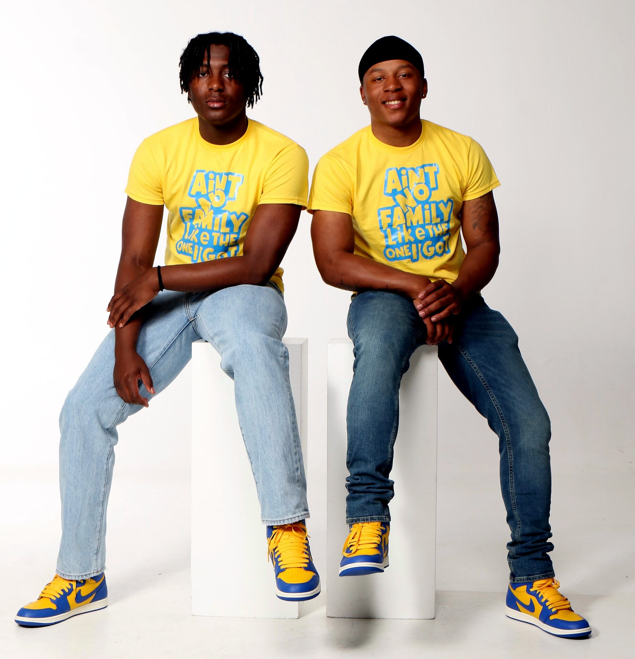 Two men in matching yellow T‑shirts sit on white blocks, wearing blue and yellow sneakers; shirt text reads 'Ain't No Family Like The One I Got'.