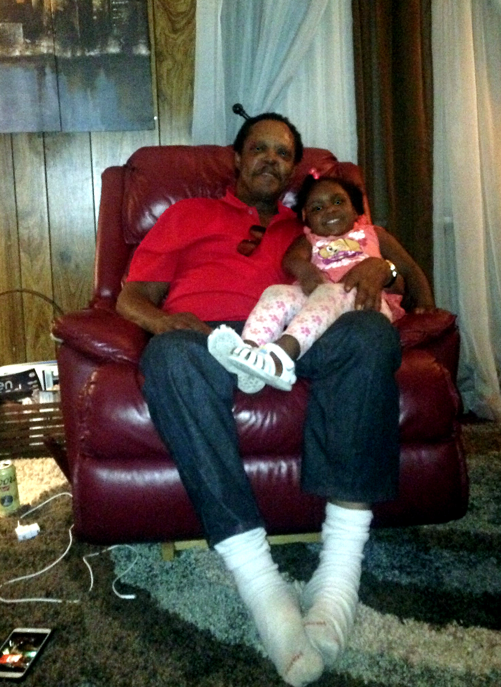 Elderly man in a red polo sits in a red leather chair with a smiling little girl on his lap in a cozy living room.