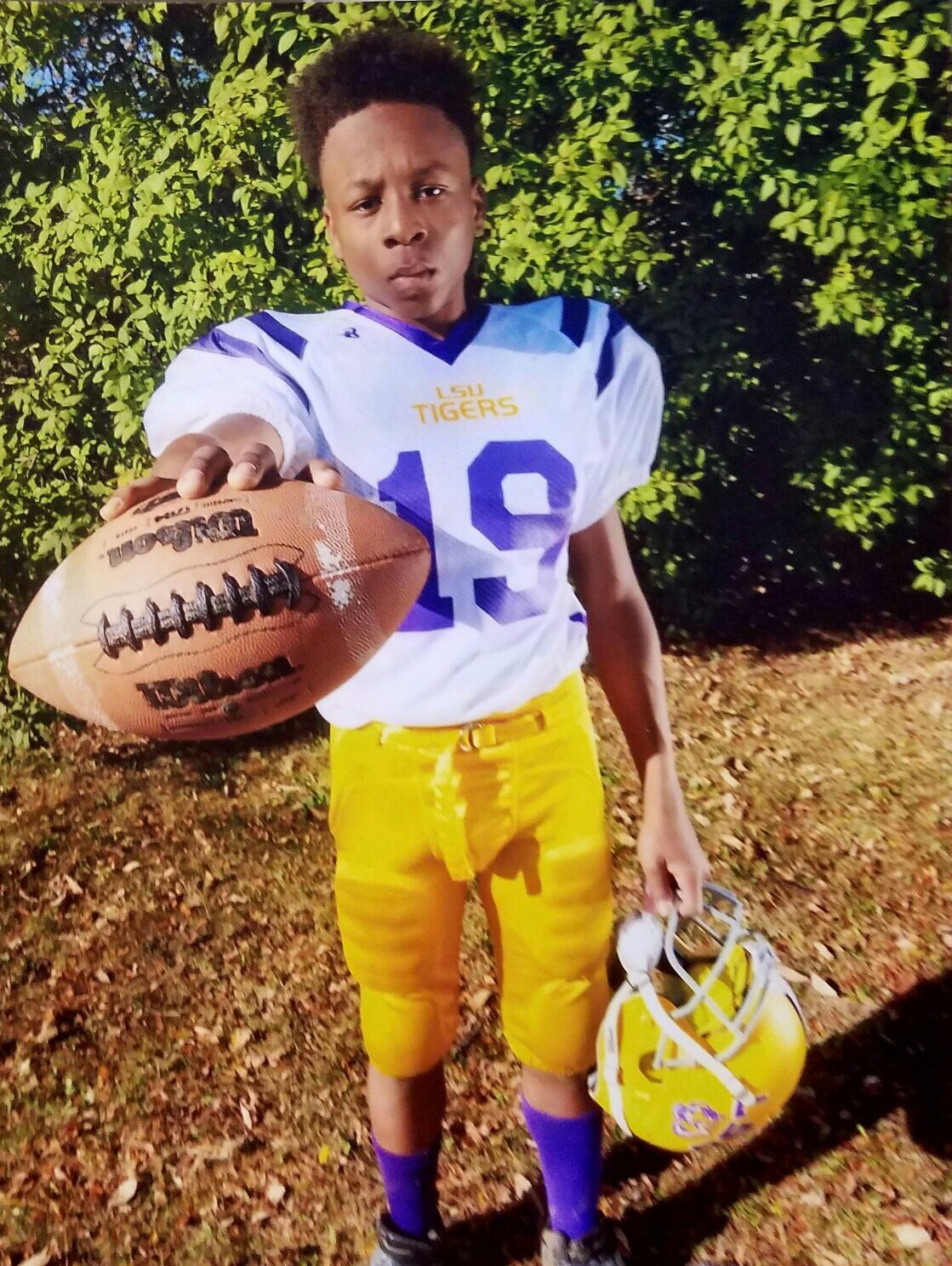 Young football player in LSU Tigers jersey (#19) extends a football toward the camera outdoors, with a yellow helmet in hand behind him.