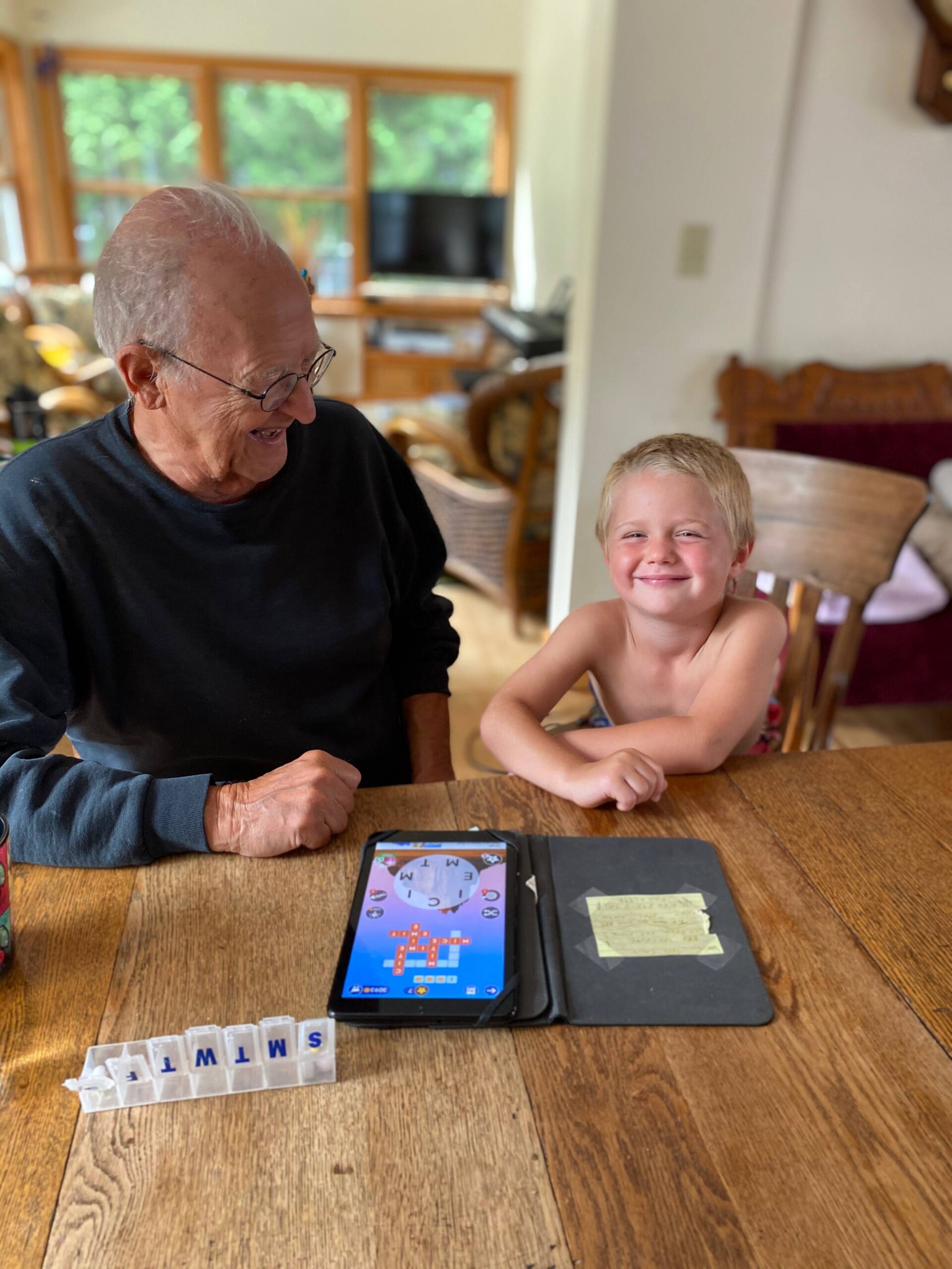 Grandfather and young boy smile at a tablet game on a wooden table, with letter tiles laid out in a rack nearby.
