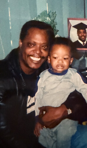 Man holding a smiling toddler indoors with a graduation photo on the wall behind them.