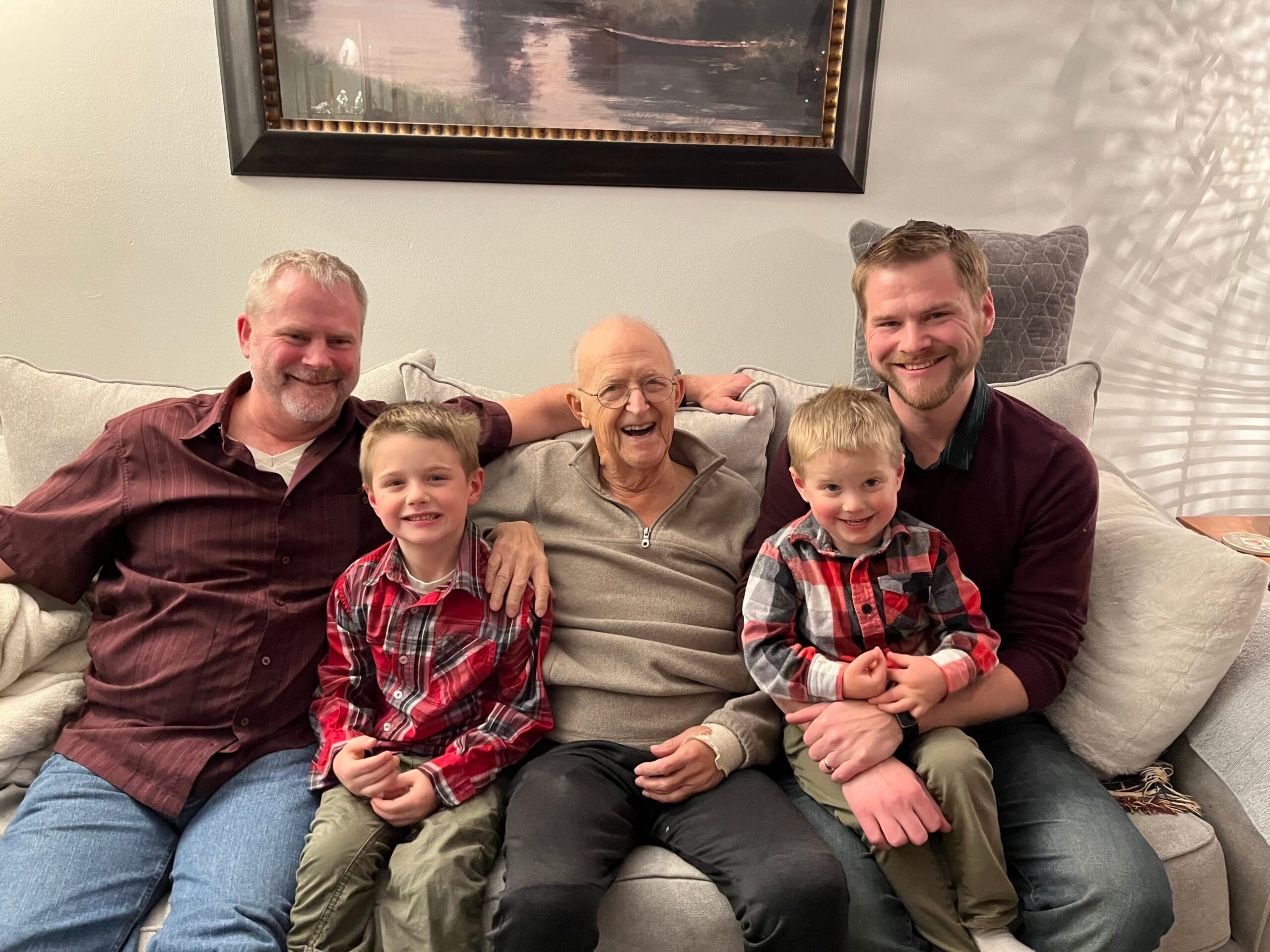Five people sit closely on a light gray sofa, with an elderly man in the center, two young boys in plaid shirts, and two men on the ends, all smiling at the camera.