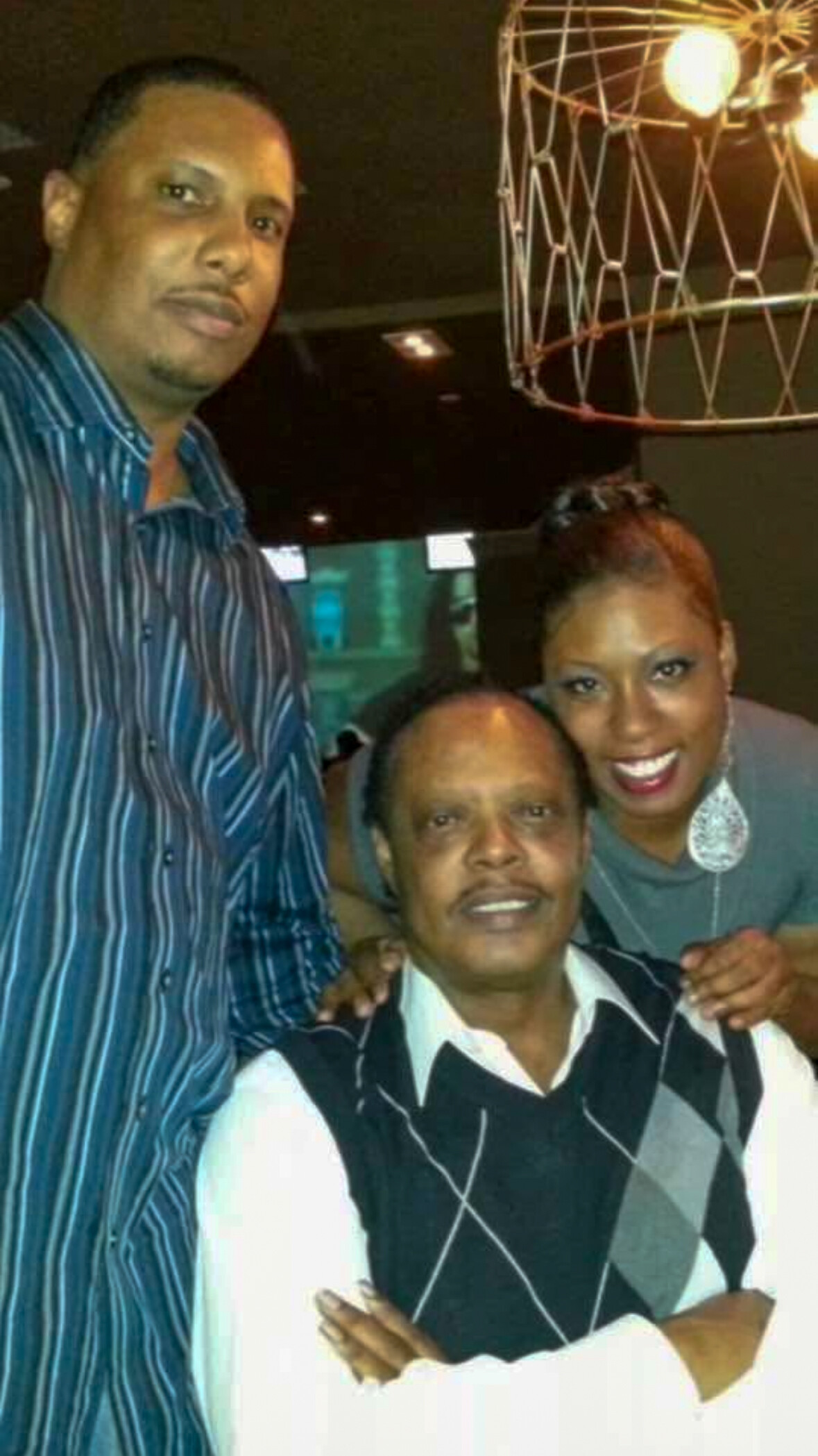 Three adults posing together for a photo indoors: a tall man in a blue striped shirt on the left, a woman in a gray top with large earrings on the right, and a seated older man in front wearing an argyle vest, all smiling.