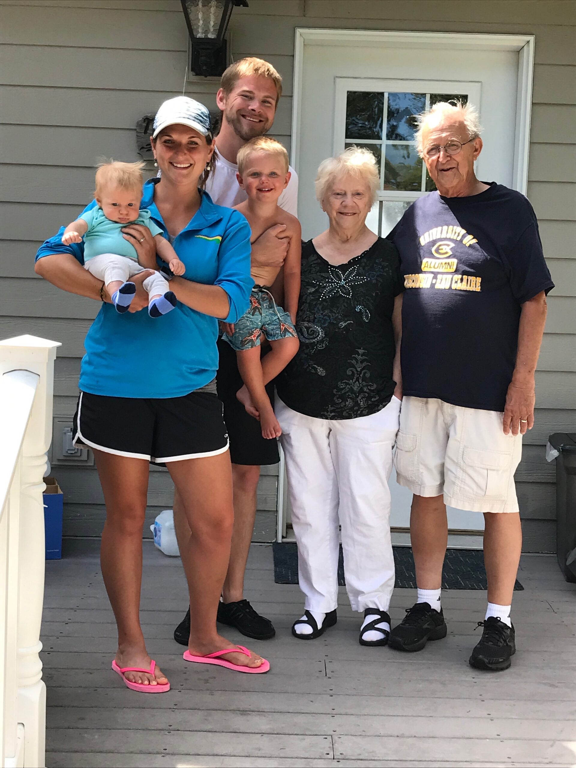 Family group photo on a front porch: woman holding a baby, man standing behind with a young boy, and two elderly grandparents smiling beside them.