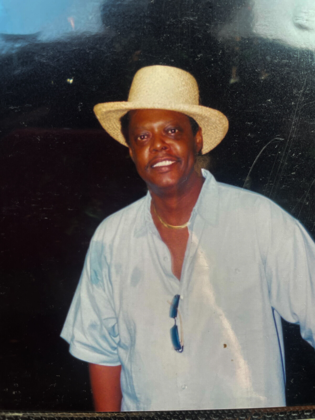 Man wearing a straw hat and light blue button-up shirt, smiling at the camera; sunglasses hang from his shirt and a gold necklace is visible (outdoor portrait).