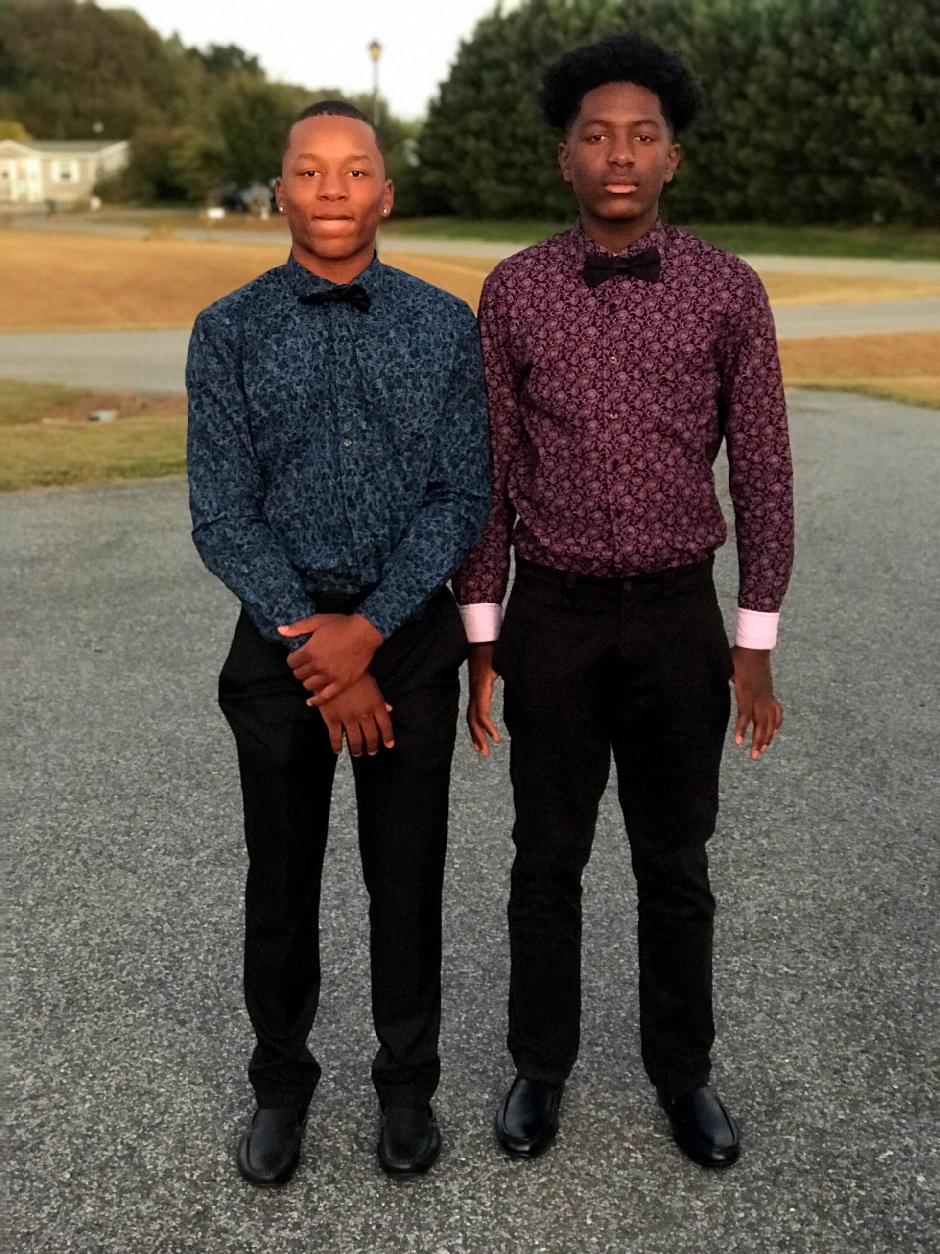 Two young men stand side by side on a gravel road, wearing patterned dress shirts with bow ties and black pants.