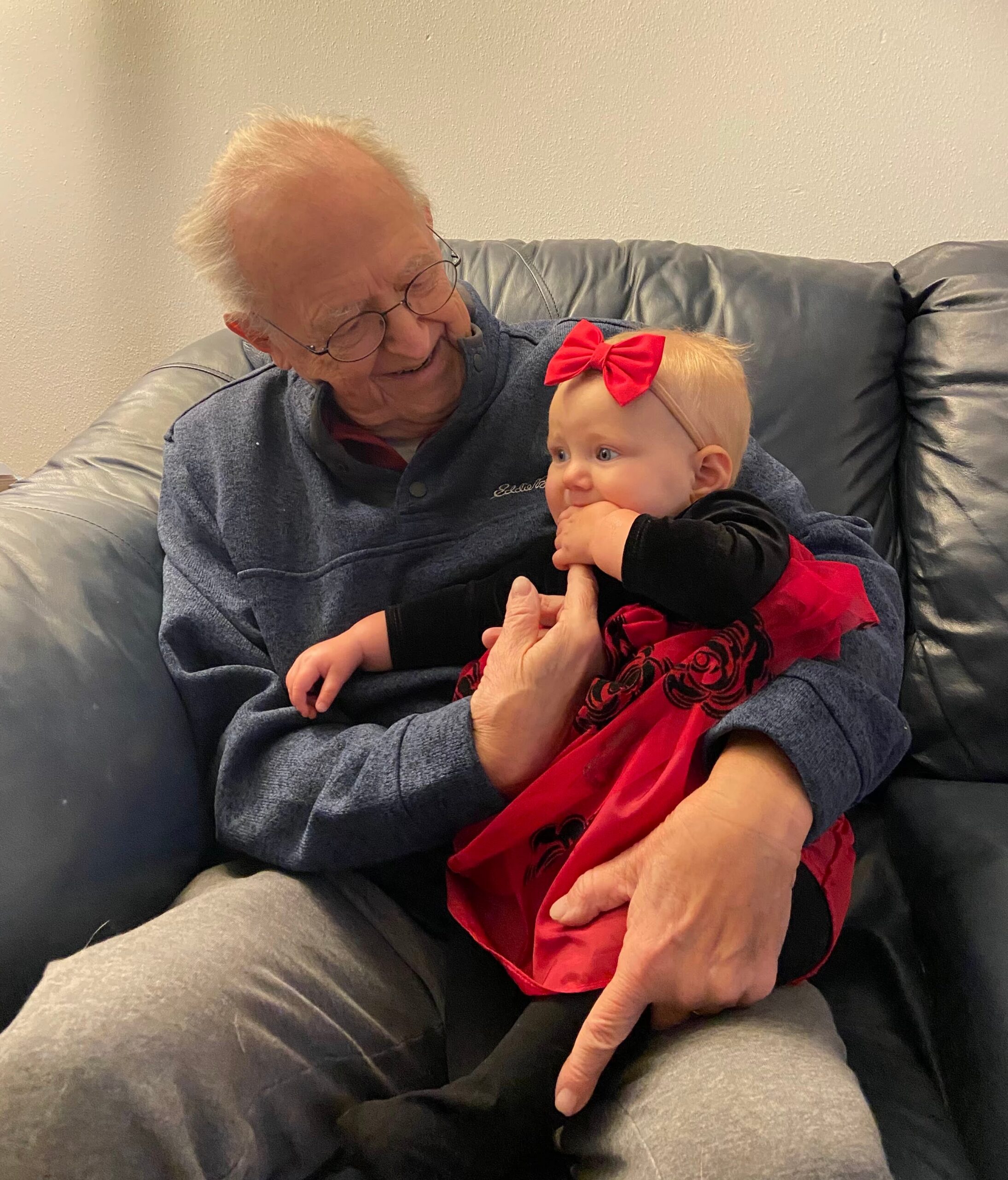 Older man with glasses sits on a leather couch, smiling at a baby girl in a red dress with a bow in her hair held on his lap