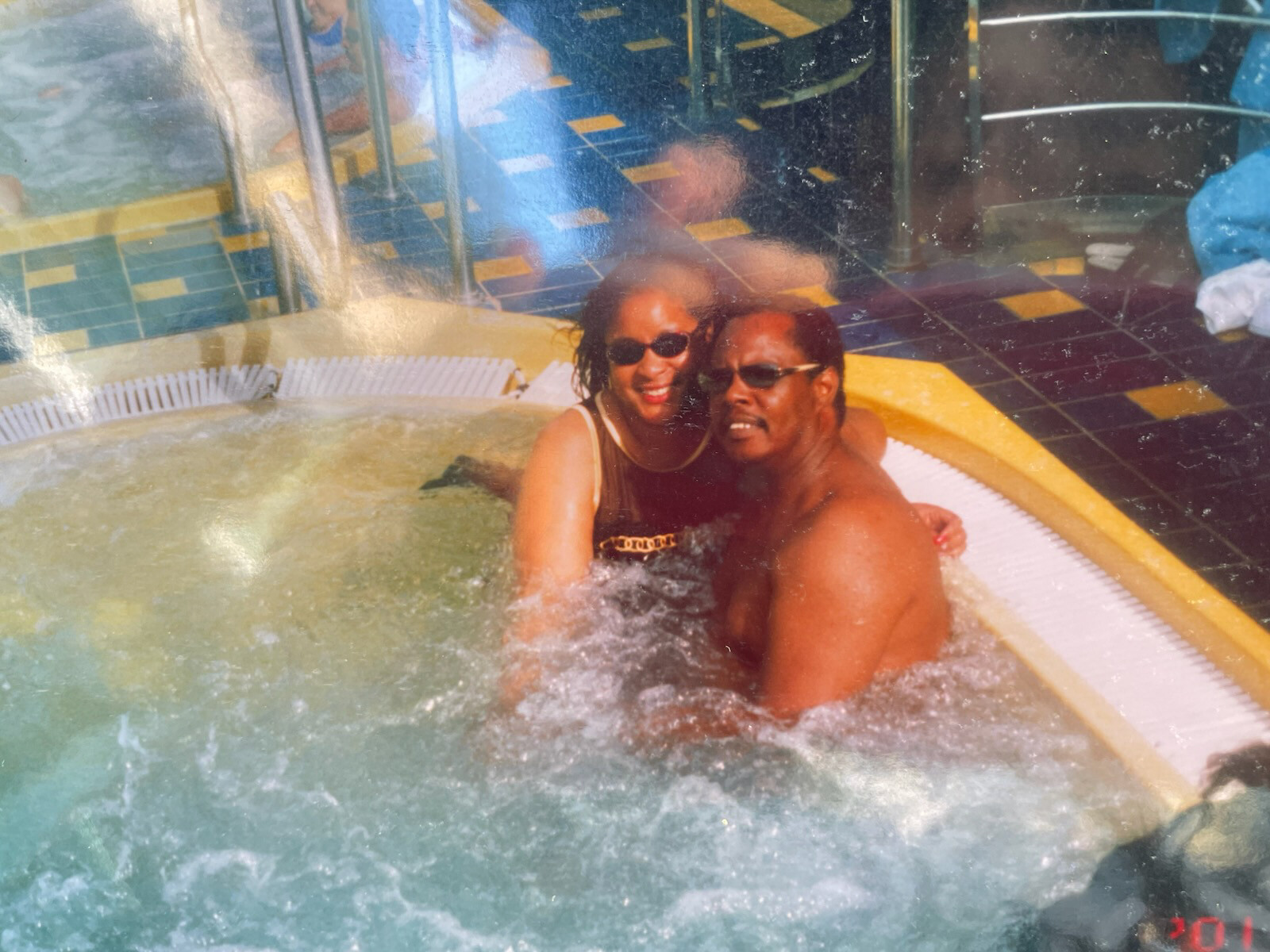 Couple wearing sunglasses smiles and hugs in a circular hot tub indoors with colorful tiled floor nearby, camera snapshot style