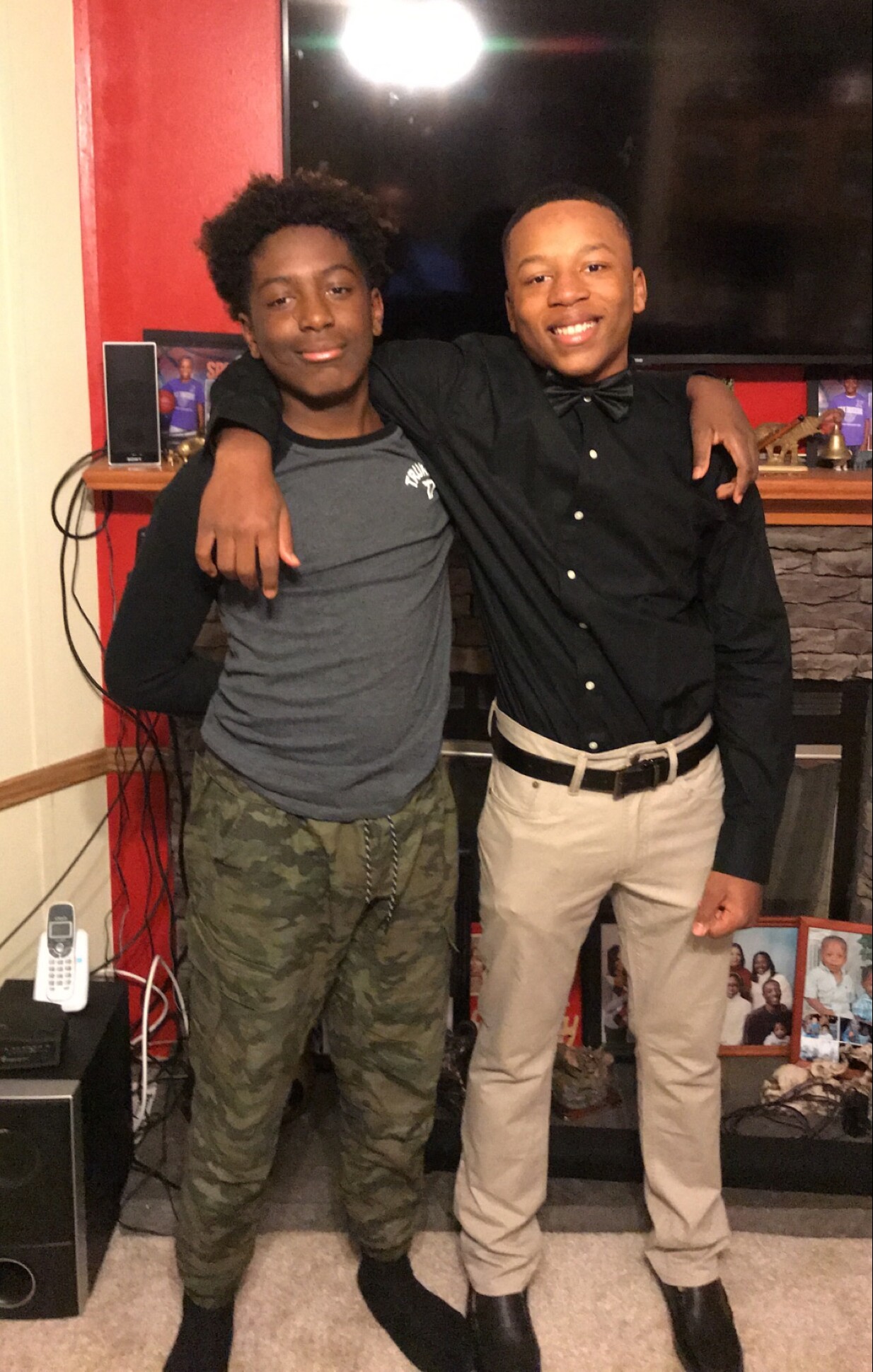 Two young men pose with arms around each other in a living room, smiling at the camera.
