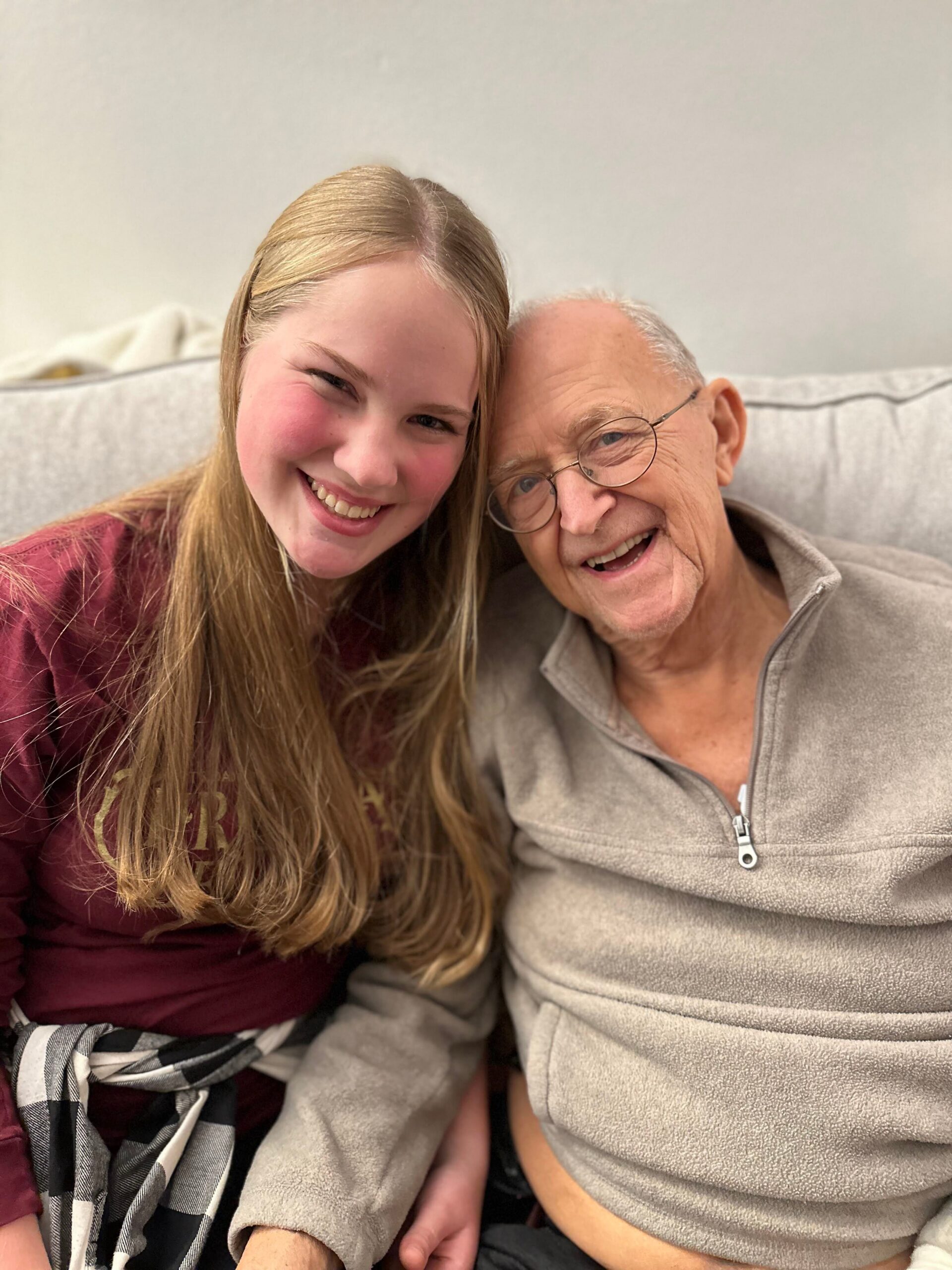 Young woman and an elderly man smiling warmly, sitting close on a couch for a selfie.