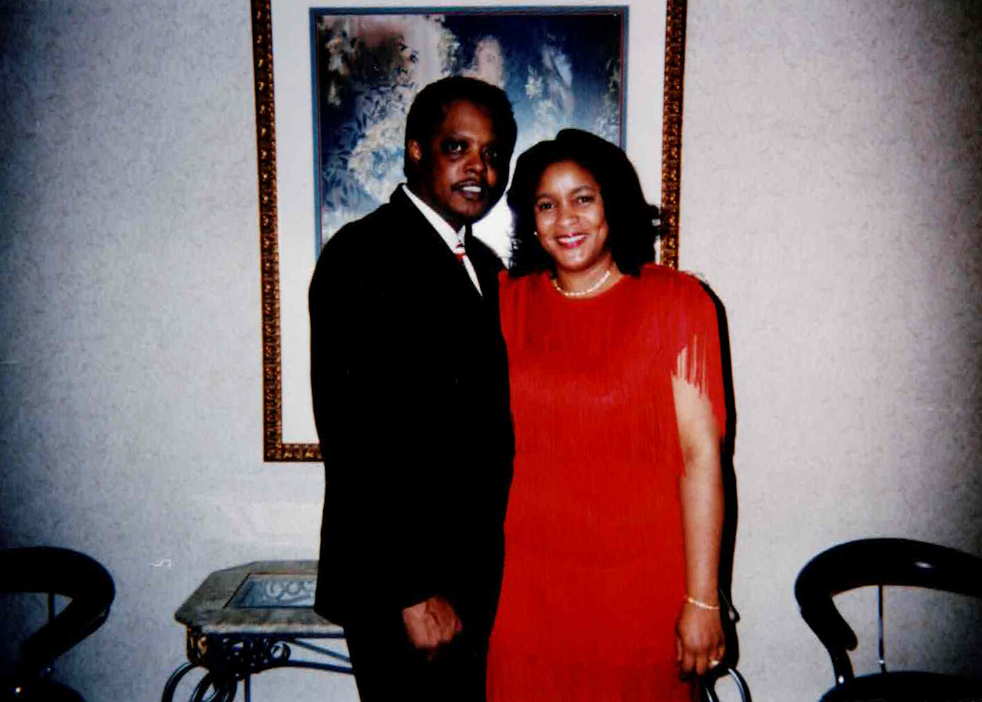 Man in a black suit and woman in a red dress pose together indoors, smiling in front of a framed painting.