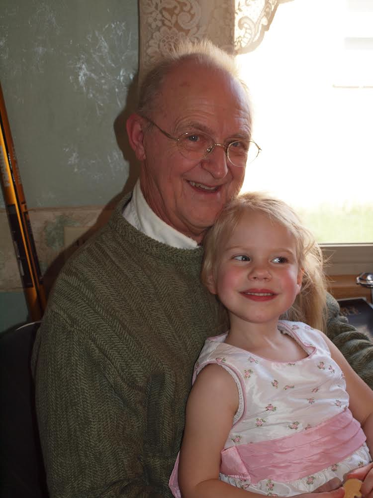 Grandfather with glasses smiling while sitting with a young girl on his lap near a sunlit window.