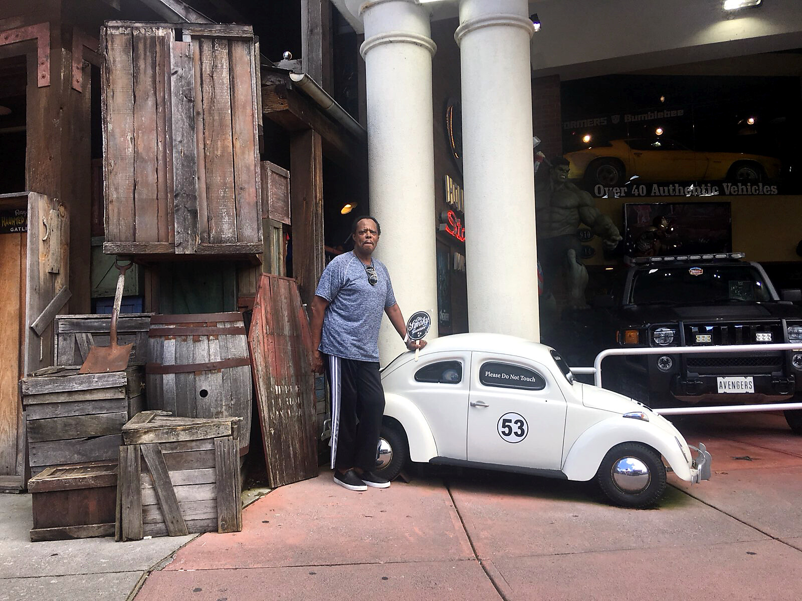 Man stands next to a white vintage Volkswagen Beetle labeled No. 53 with a 'Please Do Not Touch' sign on its window outside a car showroom with wooden crates nearby.