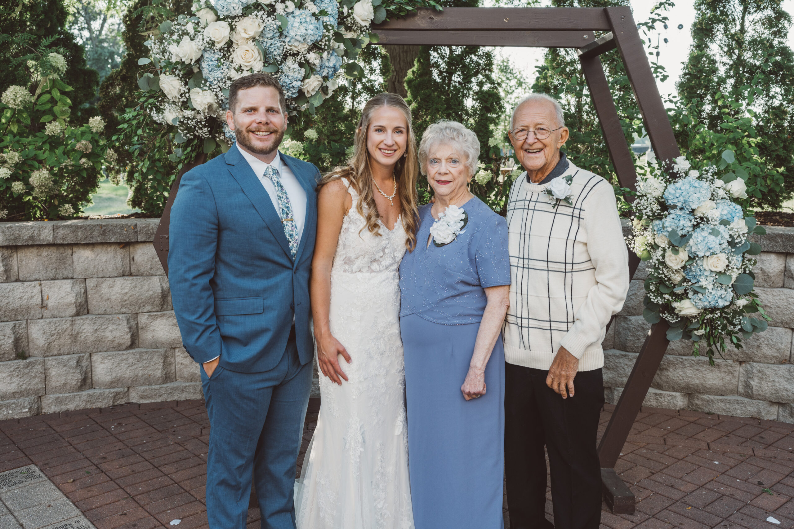 Bride and groom with two older relatives posing at a wedding beneath a floral arch.