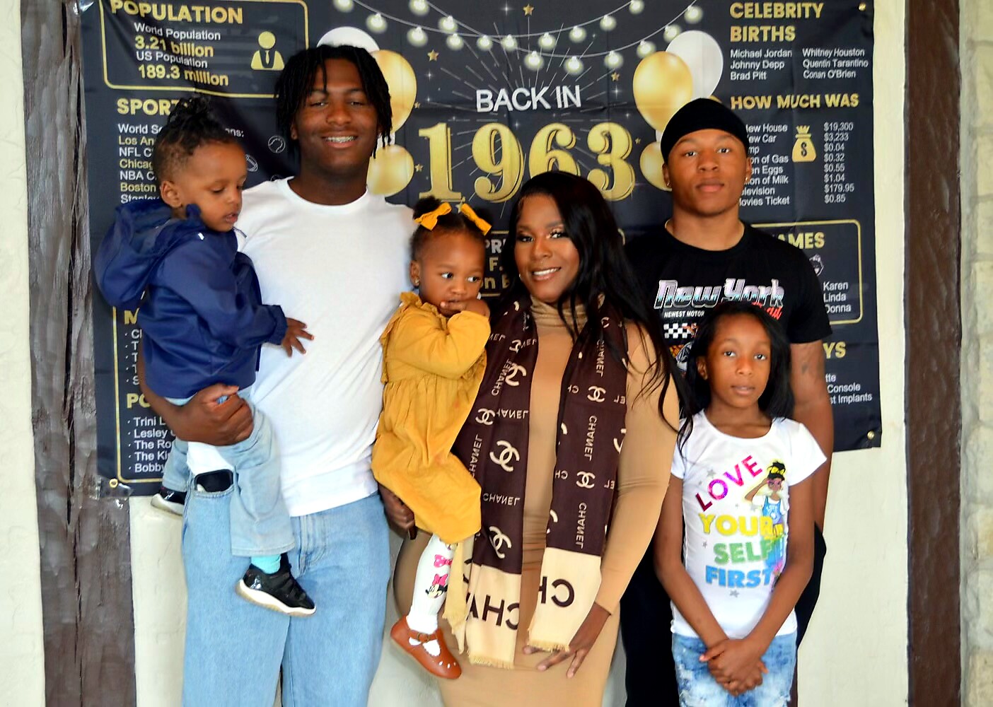 Six-family portrait in front of a celebratory poster reading 'Back in 1963' with balloons in the background, everyone smiling. Two children are being held by adults.