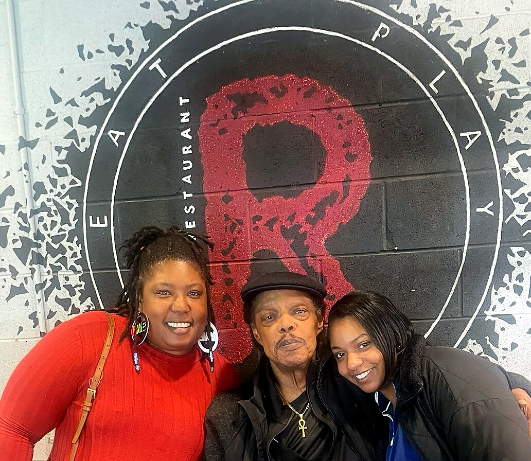 Three women smiling and posing in front of a black circular mural with a red abstract design.