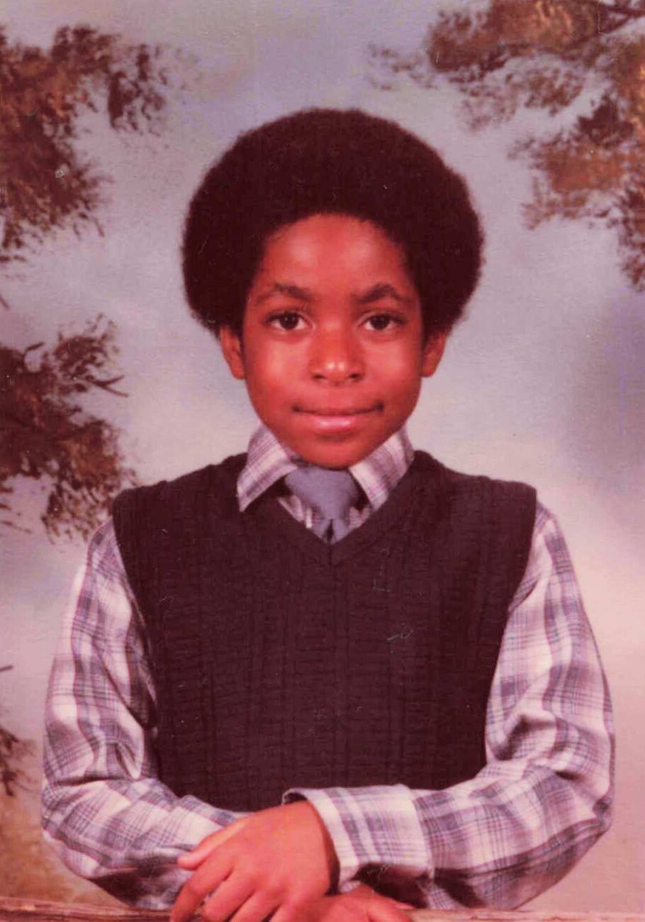 Portrait of a young boy with a large afro, wearing a plaid shirt, tie, and dark sweater vest for a school photo.