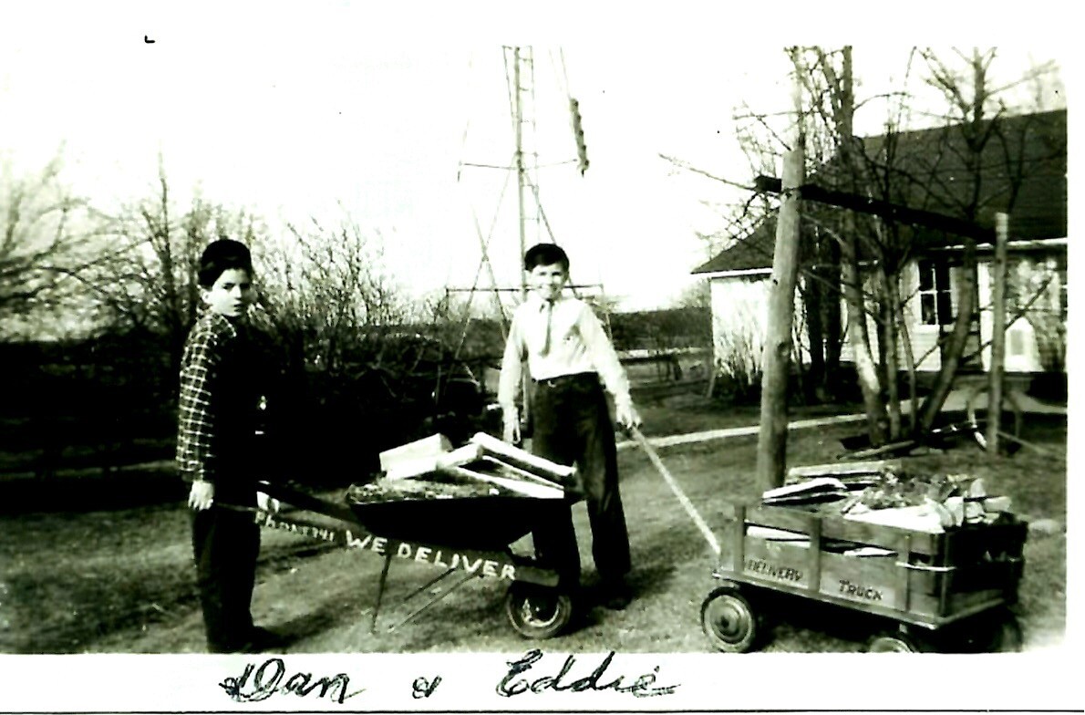 Two boys stand by a wheelbarrow labeled We Deliver in a yard, with a smaller cart loaded with goods nearby.