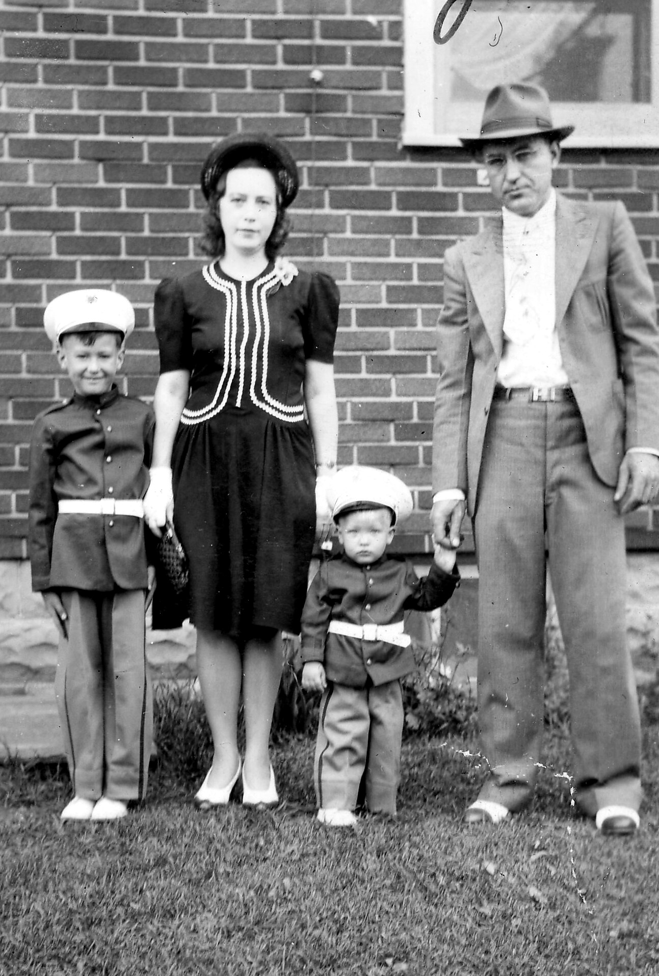 Family of four posing in front of a brick wall: mother in a black dress with white trim and hat, father in a suit and fedora, and two young boys in matching uniforms with hats, holding hands outside the house.